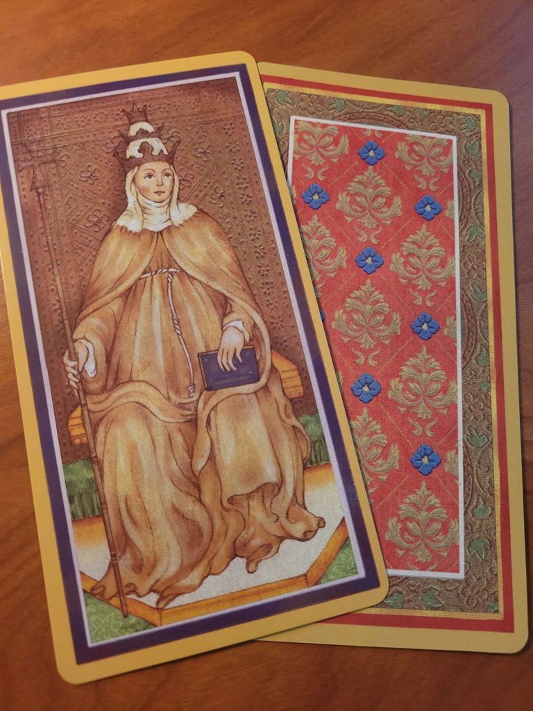 Image of the Popess card wearing the triple crown and holding a staff with the back of another card from this deck. 